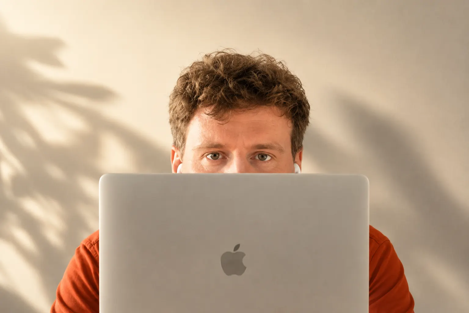 Chris looking over the top of his laptop, in a burnt orange shirt against a warm sunlit wall